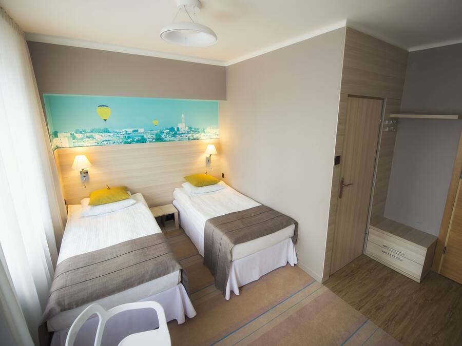 A hotel room with two single beds, a painting of a cityscape, a wooden wardrobe, and a shower area.