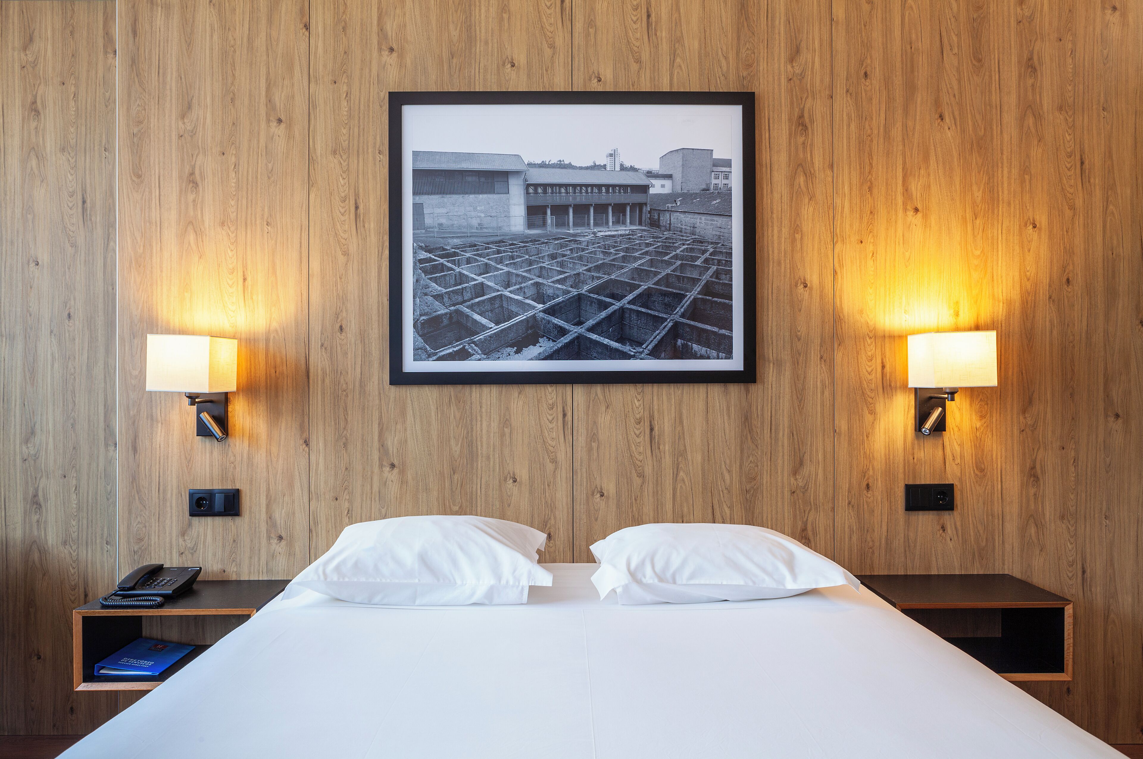 A hotel room with a bed, two bedside lamps, a framed picture on the wall, and a nightstand with a phone and book.