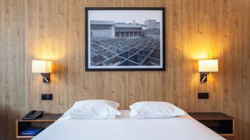 A hotel room with a bed, two bedside lamps, a framed picture on the wall, and a nightstand with a phone and book.