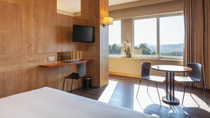 Une chambre d’hôtel équipée d’un lit, d’un bureau, de deux chaises, d’une télévision et d’une grande fenêtre donnant sur un paysage agréable.