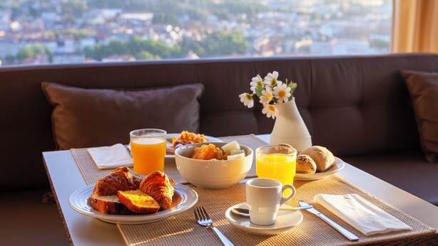 Une table de petit-déjeuner avec des croissants, un saladier de fruits et du jus d’orange, installée sur un balcon offrant une vue sur la ville.