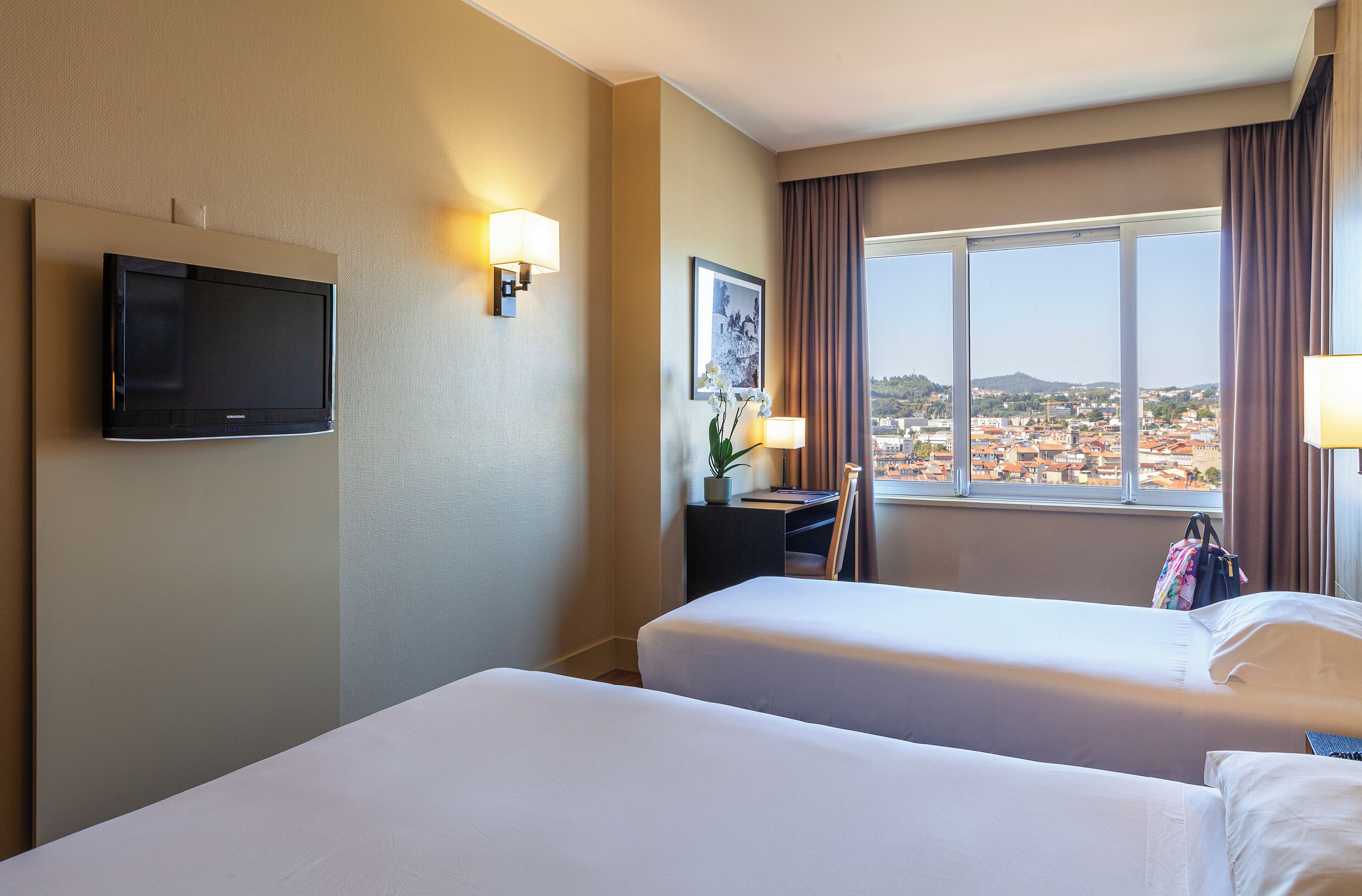 A hotel room with two beds, a TV, a desk with a lamp, a framed picture, and a view of a cityscape.