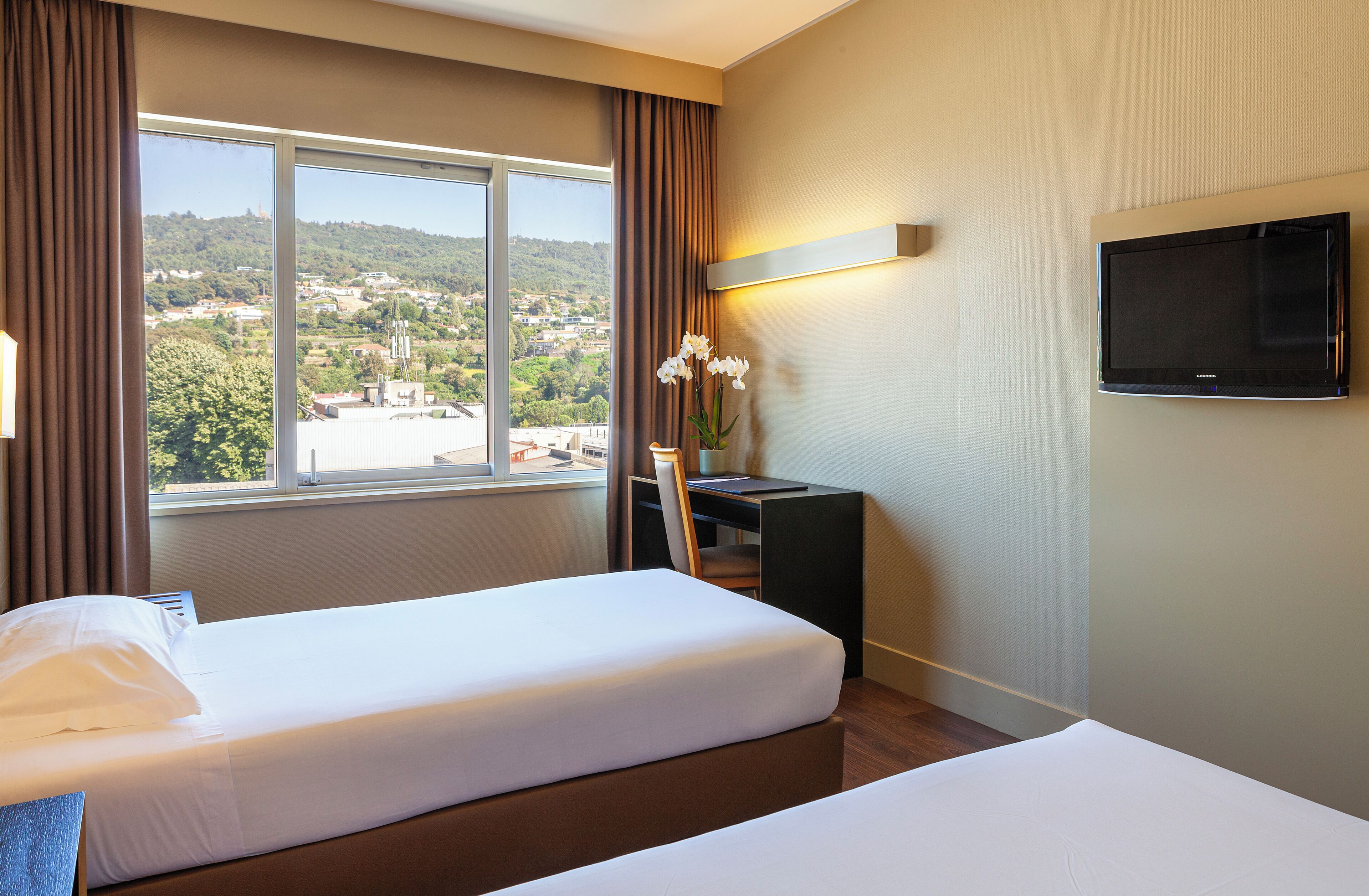 A hotel room with two beds, a desk, a chair, a TV, and a window with a view of a town and hills.