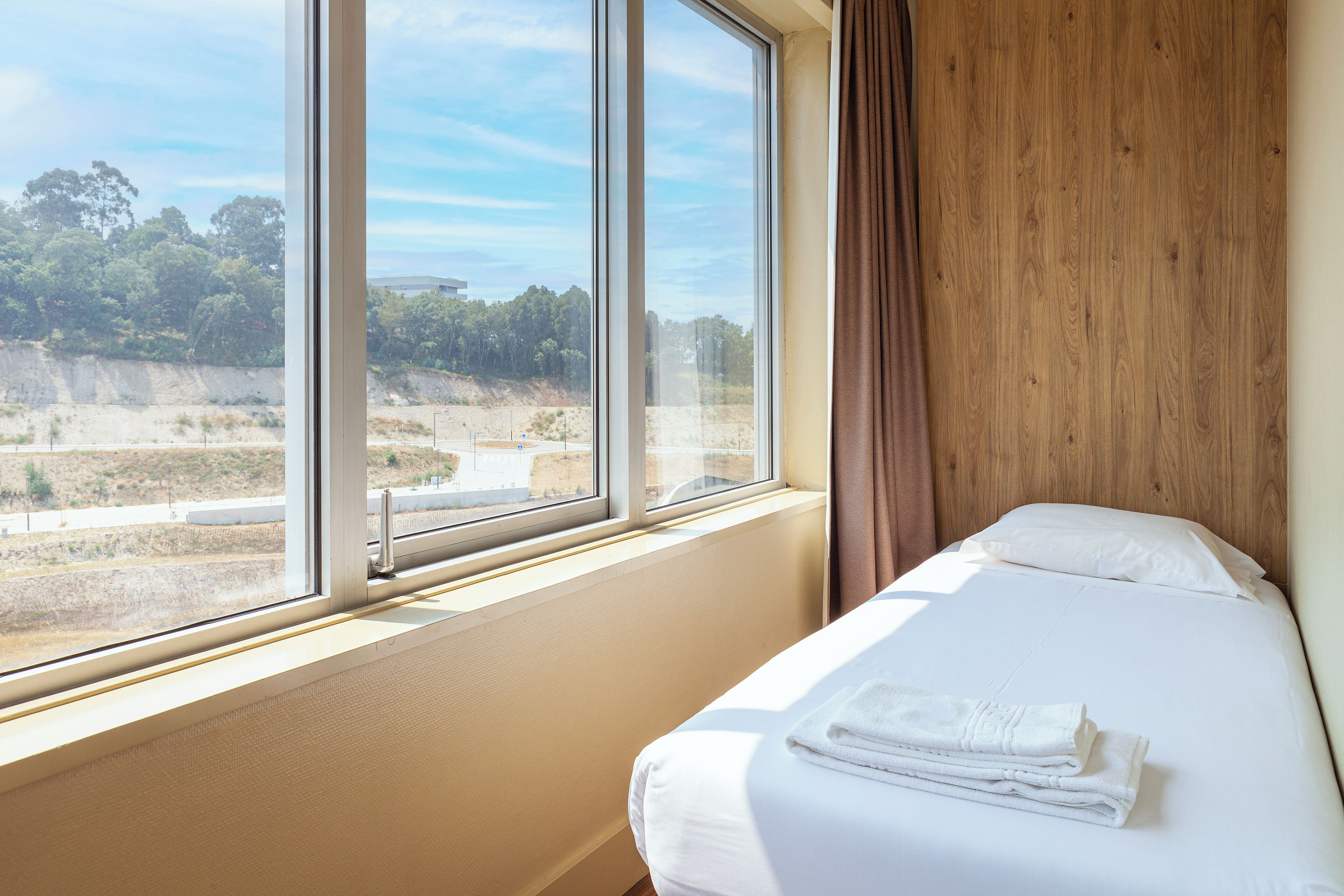 Una habitación de hotel con un ventanal grande, una cama con ropa blanca y un cabecero de madera.