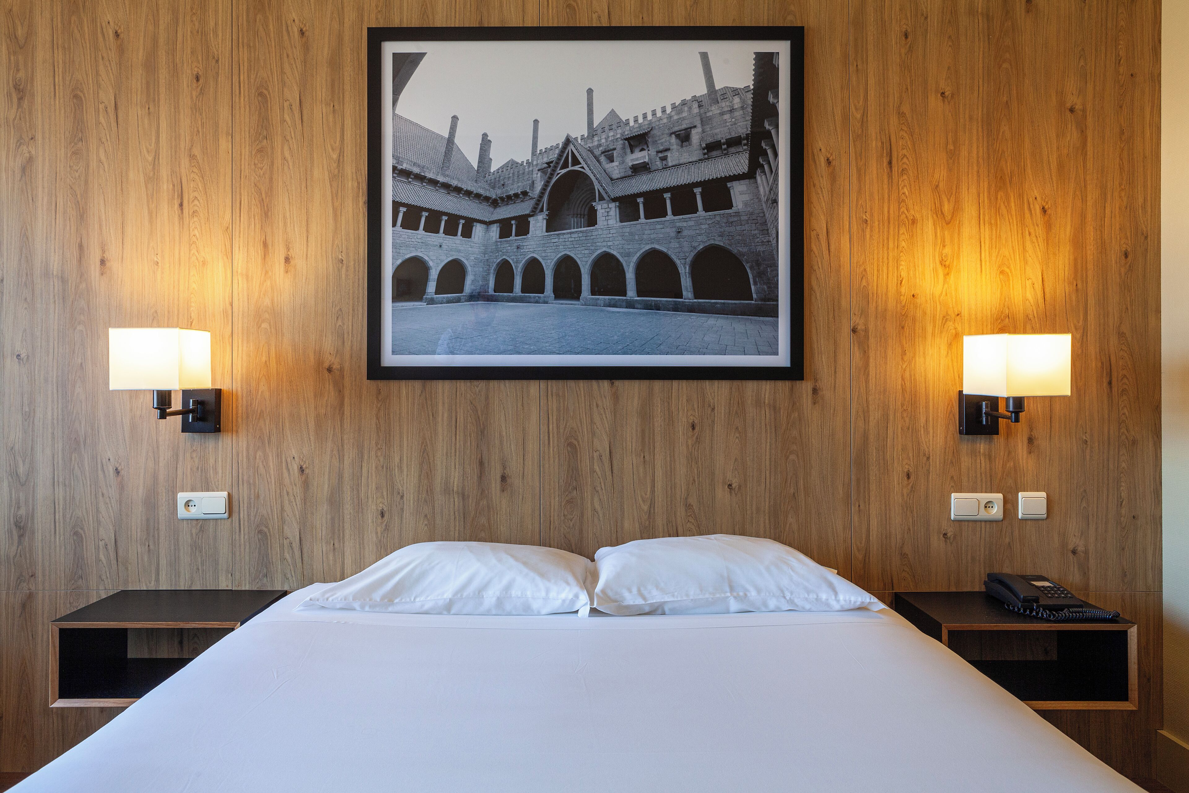 A hotel room with a large bed, two bedside tables, a framed picture on the wall, and two wall-mounted lamps.
