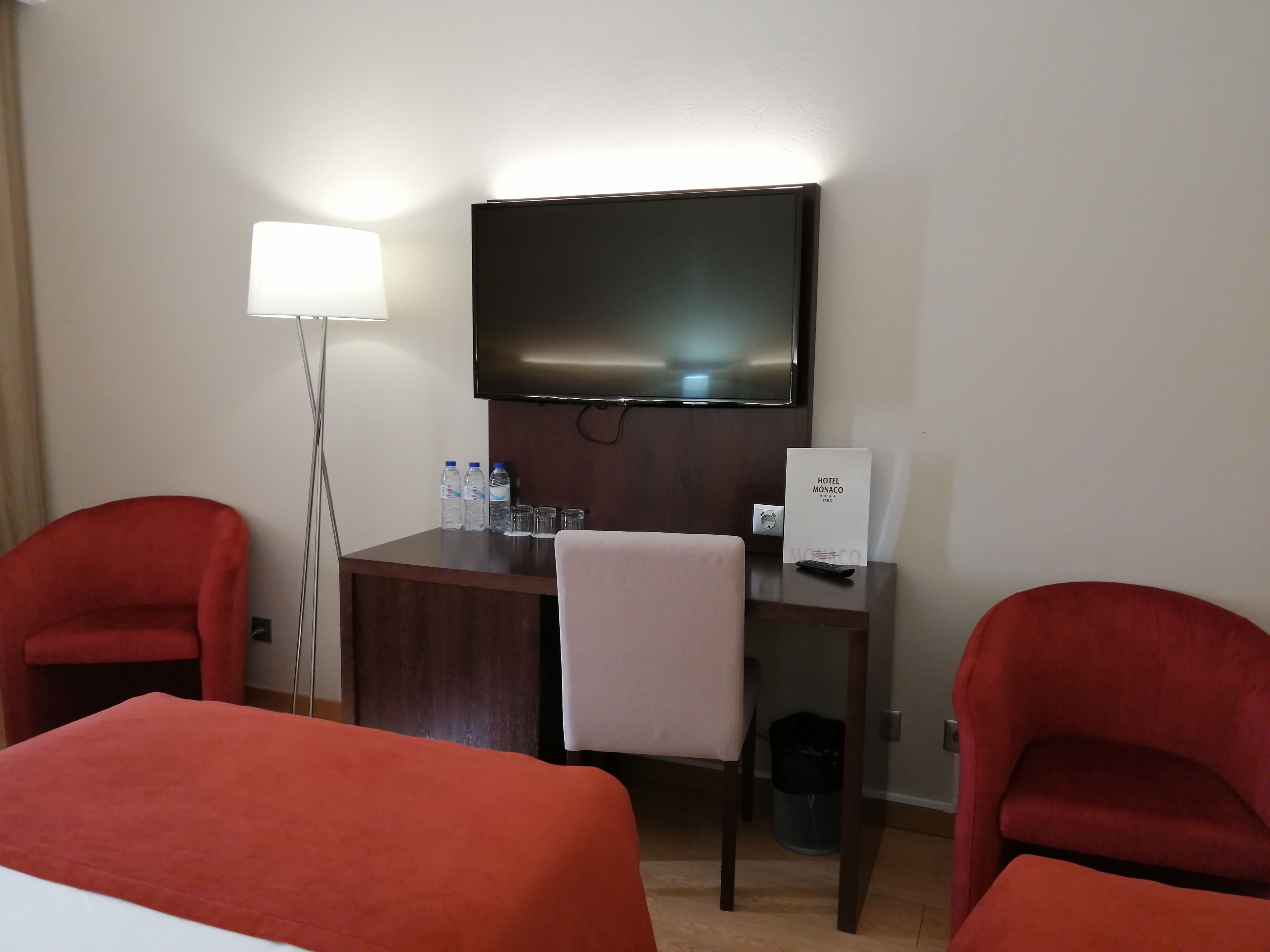 Habitación de hotel con una cama, dos sillones rojos, un escritorio con televisor de pantalla plana, una lámpara y un cartel con el nombre del hotel.