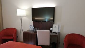 Habitación de hotel con una cama, dos sillones rojos, un escritorio con televisor de pantalla plana, una lámpara y un cartel con el nombre del hotel.