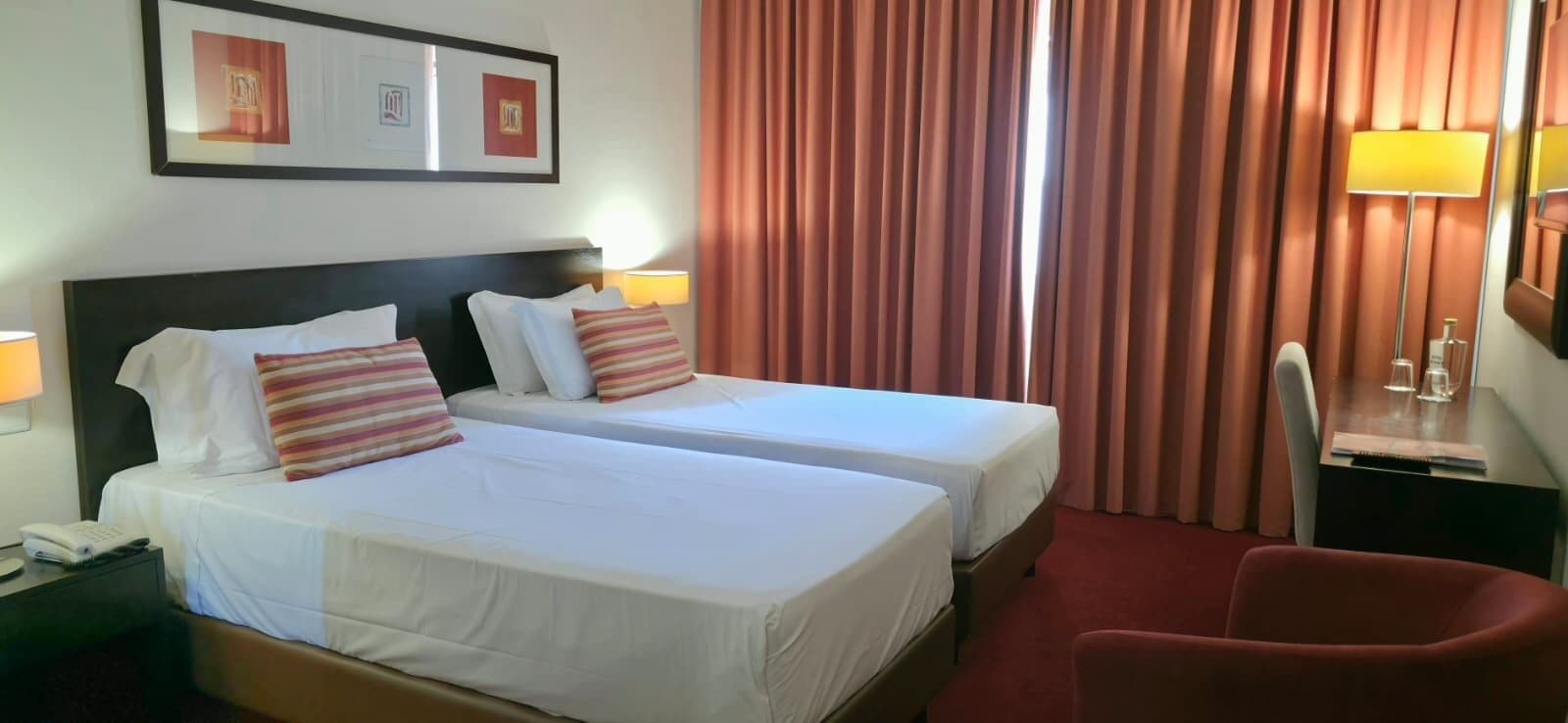 A hotel room with two beds, a desk, a chair, and a lamp.