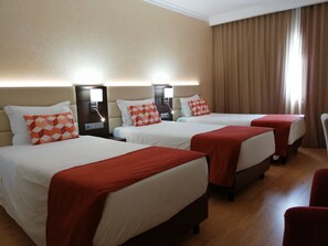 A hotel room with three beds, each with a red blanket and a patterned pillow. The room has a wooden floor and beige walls. - Hotel Mónaco (Faro)