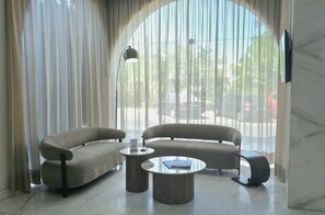A modern lounge area with two sofas and a round coffee table, large windows with sheer curtains, and a flat-screen TV mounted on the wall.