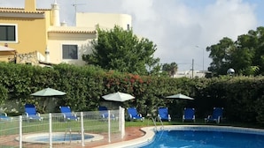 A swimming pool with lounge chairs and umbrellas, surrounded by a fence, with a building in the background.