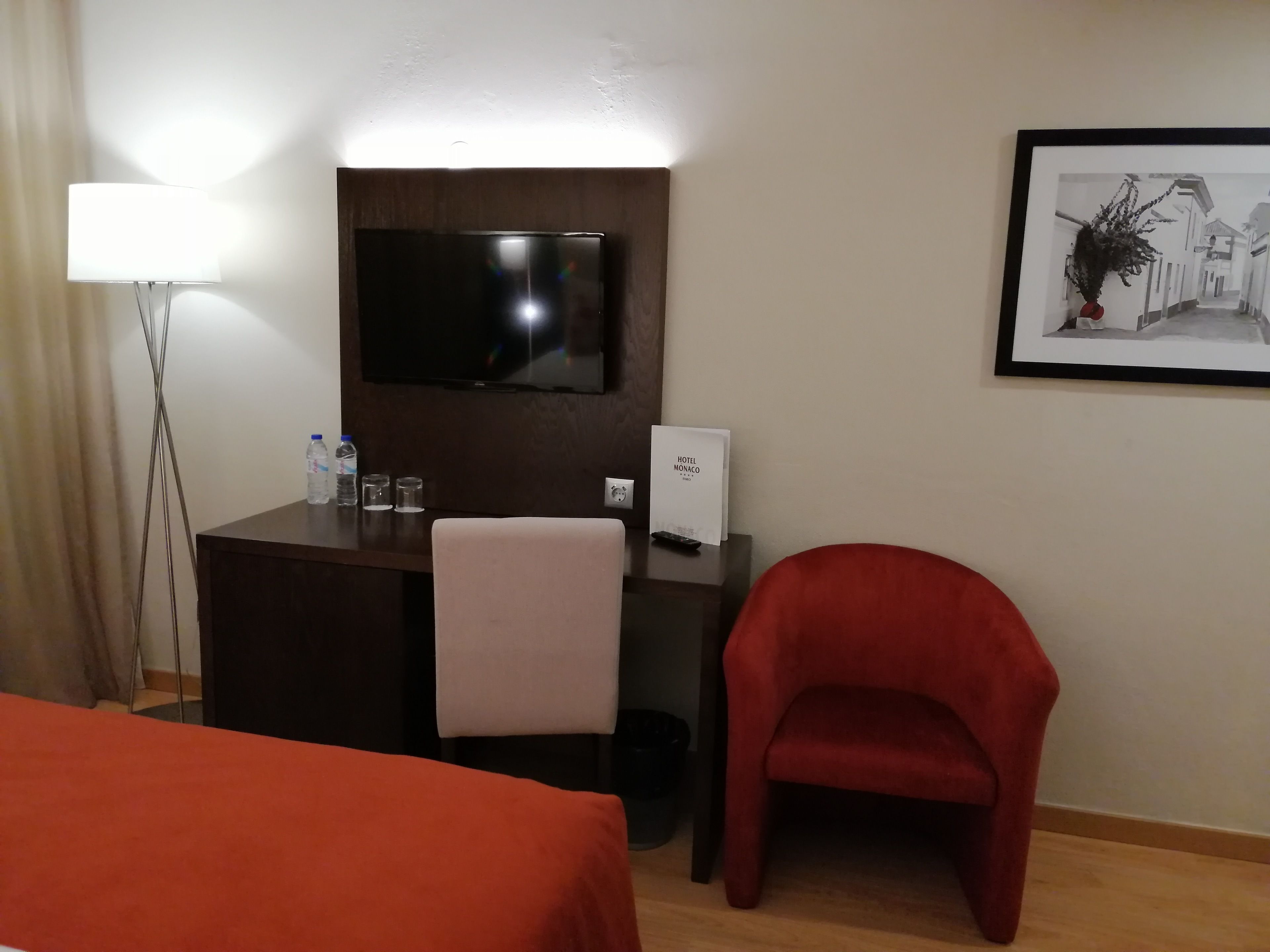 Habitación de hotel con cama, escritorio con televisor, silla roja, un cuadro en la pared y una lámpara.