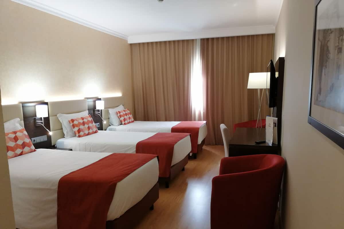 A hotel room with three beds, each with a red blanket and white bedding. There is a wooden floor, a desk with a chair, and a large window with curtains.