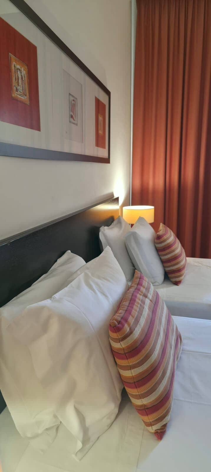 A hotel room with two beds, a headboard, a lamp, and a curtain.