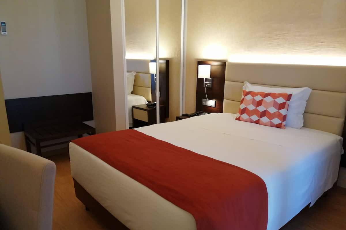 A hotel room with a large bed, a bedside table with a lamp, a chair, and a TV.