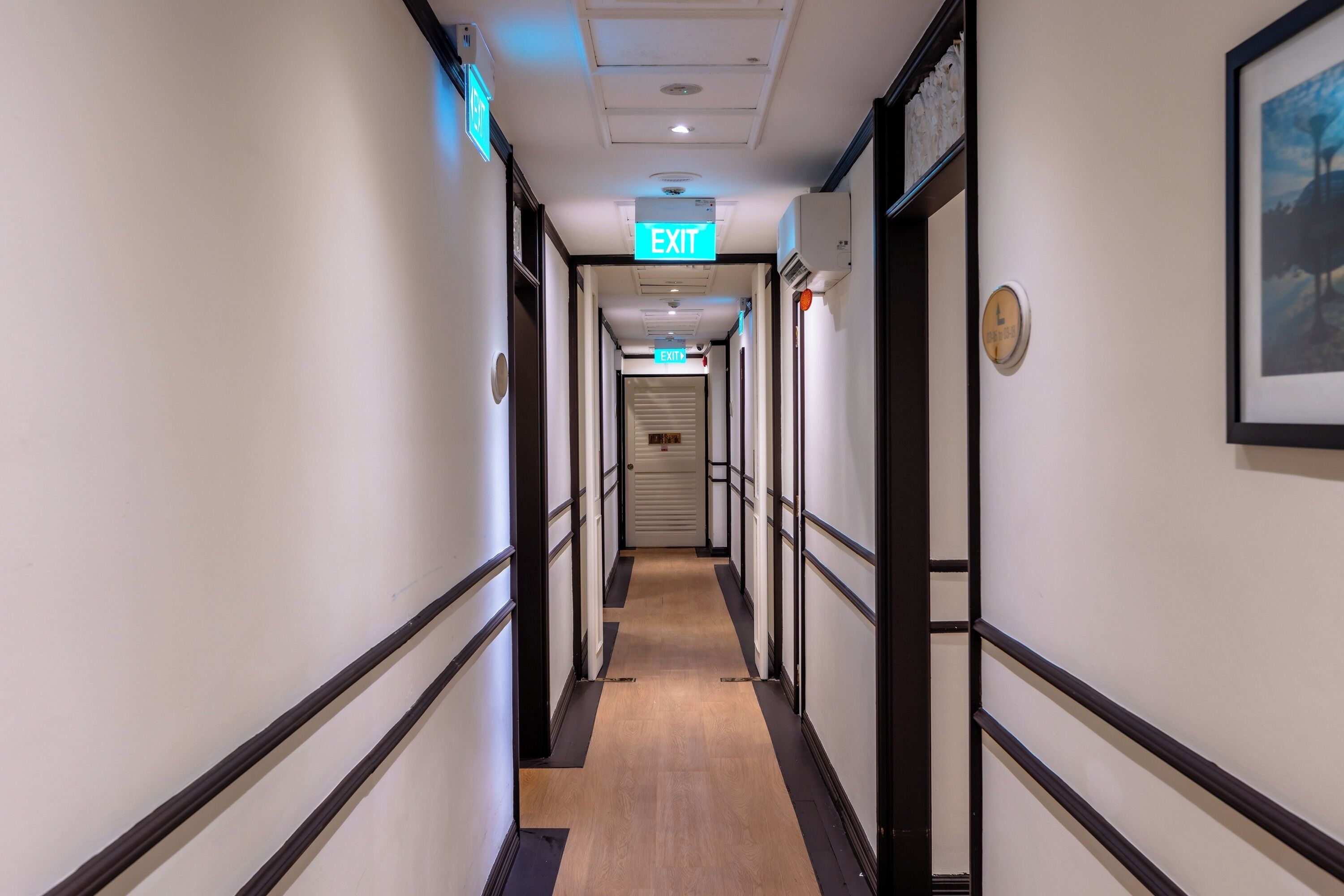 A hotel corridor with a wooden floor, white walls, and black trim. There are exit signs and a clock on the wall.