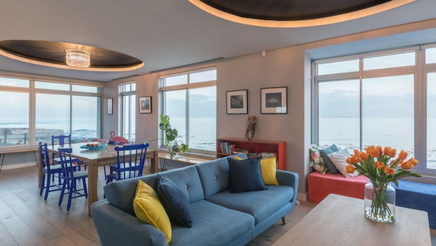 A modern living room with a blue sofa, a dining table, and chairs. There are large windows offering a view of the sea. - 24 Villa Marina (Cape Town)