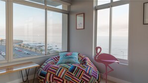 A bean bag chair with a colorful striped blanket, a pink flamingo chair, and a view of the ocean through large windows. - 24 Villa Marina (Cape Town)