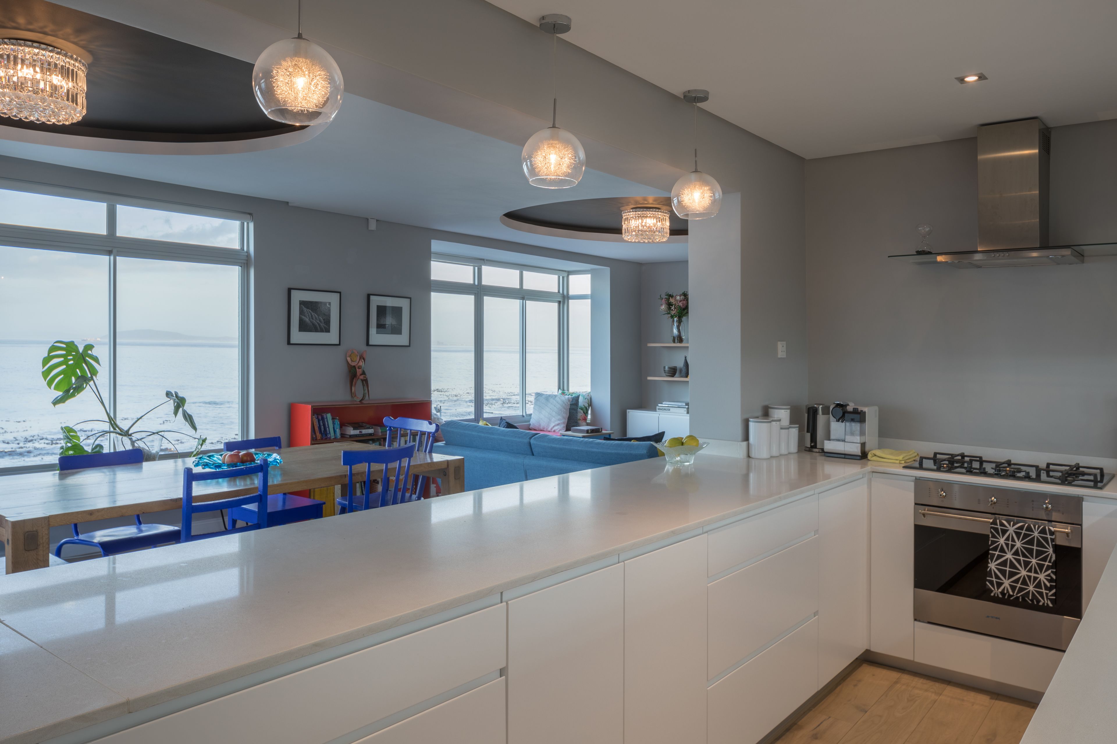 A modern kitchen with a large island, white cabinetry, and stainless steel appliances. There is a dining area with blue chairs and a view of the sea through large windows.