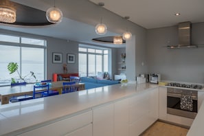 Ett modernt kök med en stor köksö, vita skåp och rostfria vitvaror. Här finns också en matplats med blå stolar och en vacker utsikt över havet genom stora fönster.
