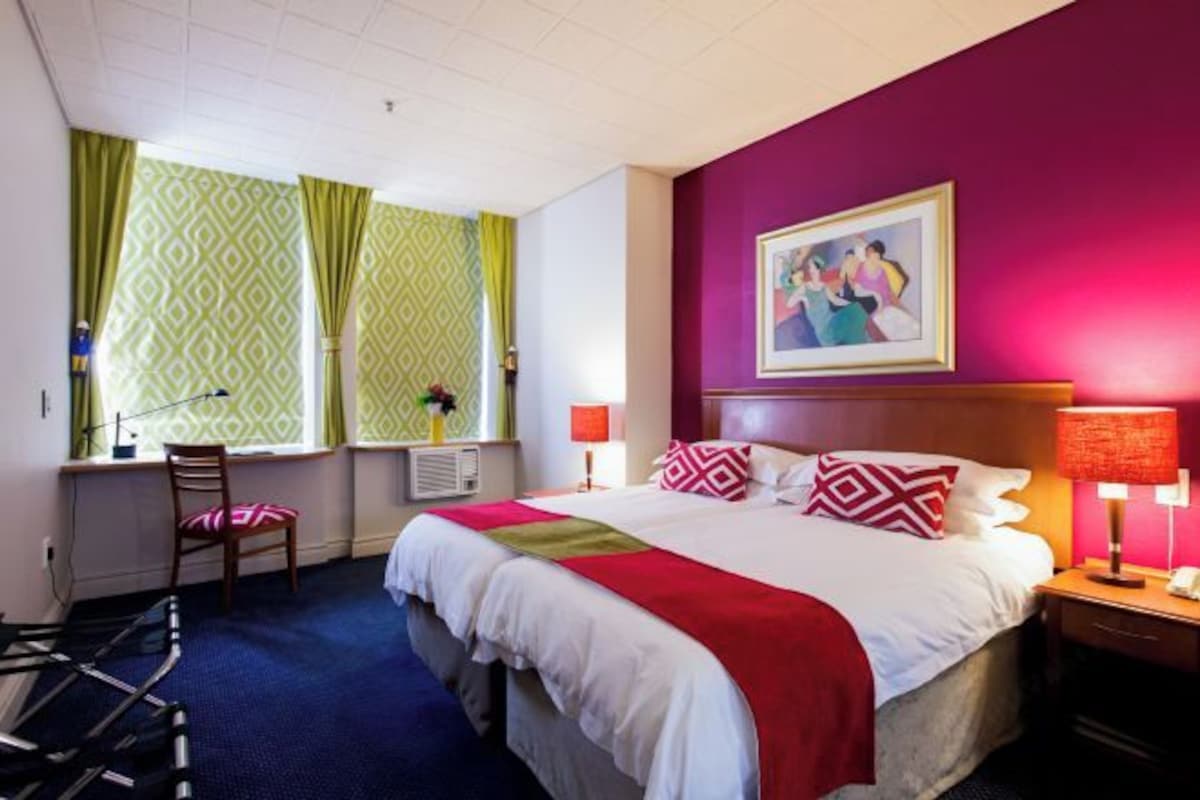 A hotel room with a large bed, a desk with a chair, a window with curtains, and a painting on the wall.