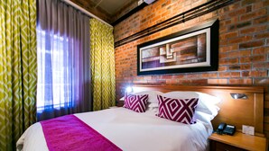 A hotel room with a brick wall, a bed with white bedding and patterned pillows, a nightstand with a phone, and a framed artwork above the bed. - Cape Town Lodge Hotel (Cape Town)