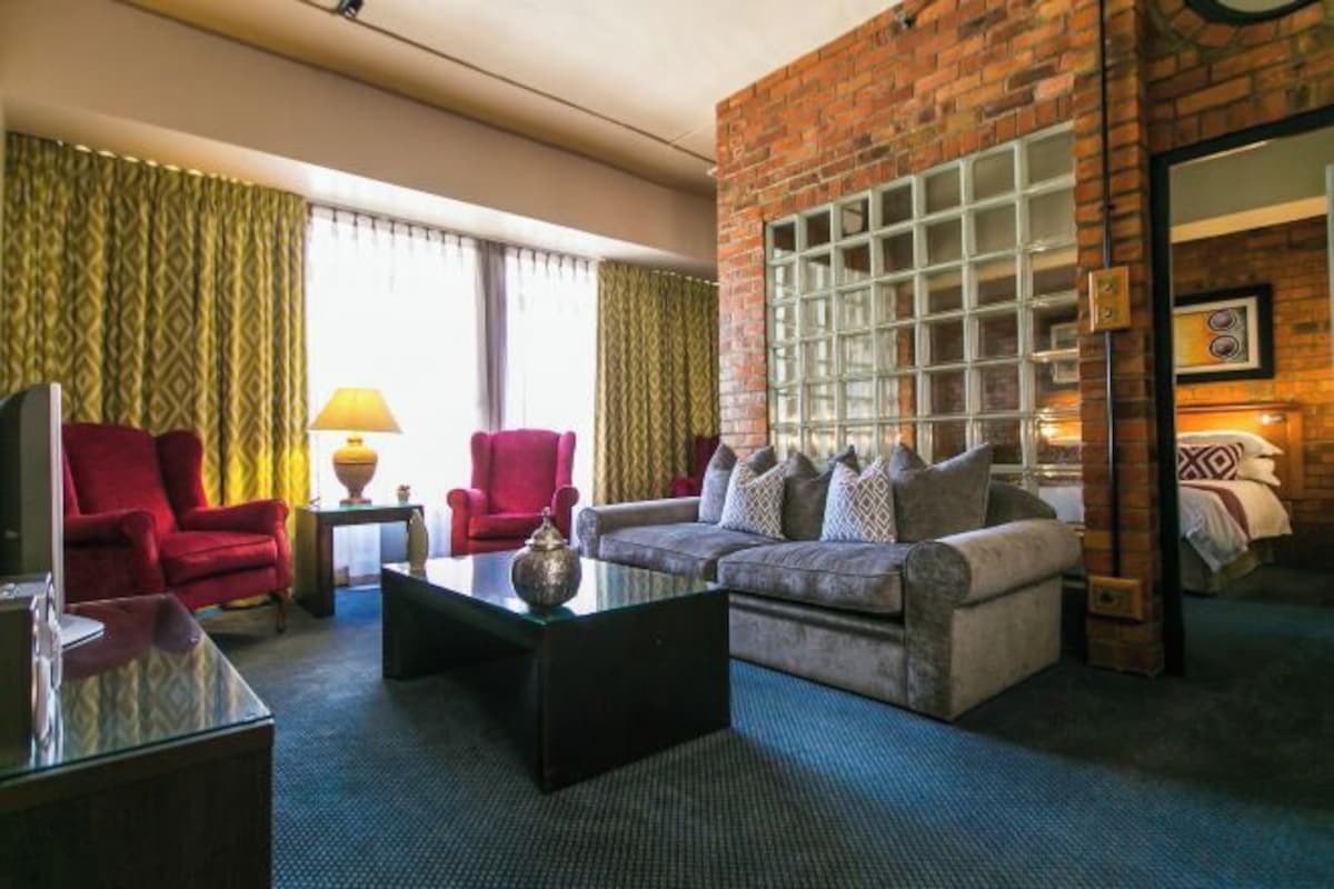 A hotel room with a brick wall, a sofa, a red armchair, a television, and a lamp.