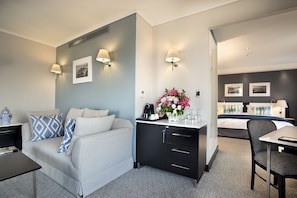 A modern hotel room with a grey sofa, a black desk, a bed with a blue bedspread, and a small dining table. - The Commodore Hotel (Cape Town)