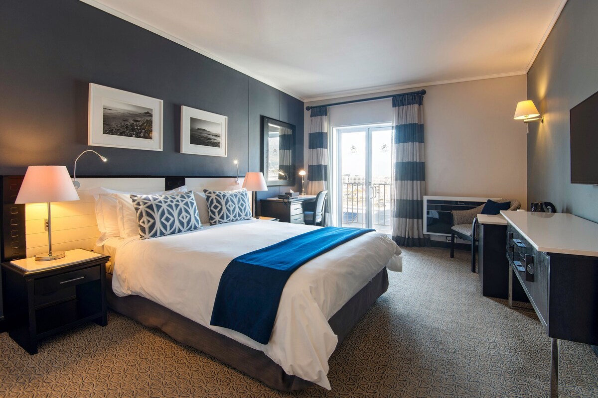 Habitación de hotel con una cama grande, mesitas de noche, un escritorio y una silla. Hay cuadros en la pared, una ventana con cortinas y un balcón visible a través de la ventana.