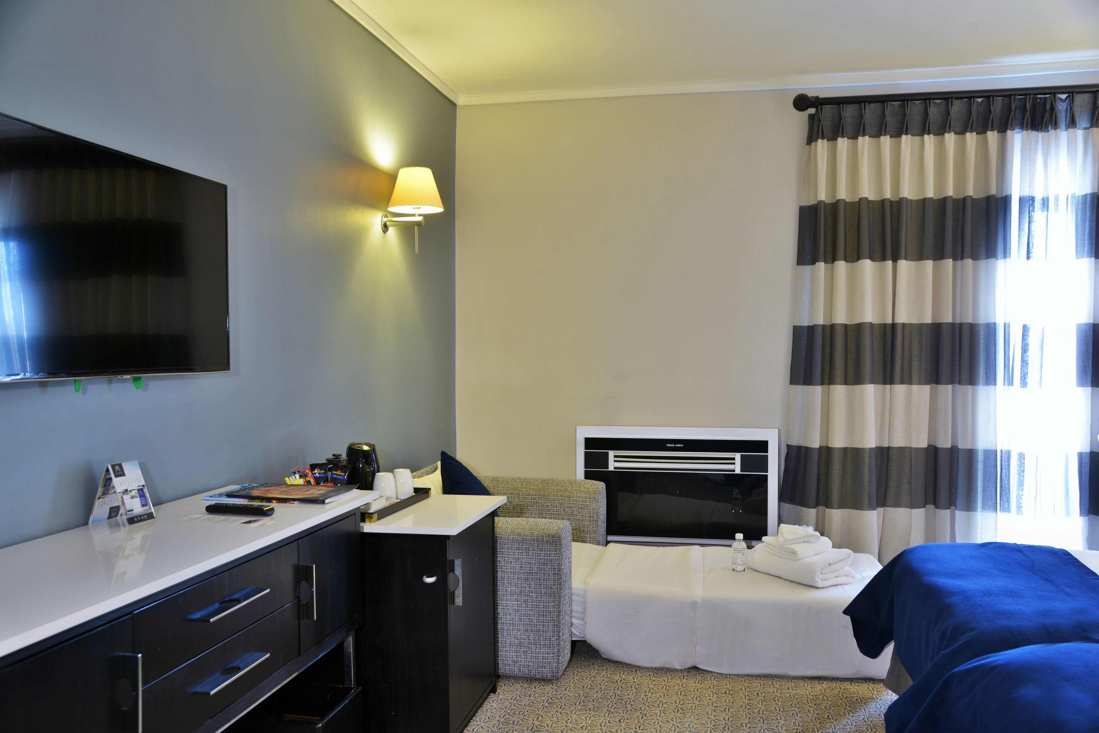 Une chambre d’hôtel équipée d’une télévision à écran plat, d’un bureau avec une lampe, d’un lit agrémenté d’un rideau à rayures et d’un petit coin salon.