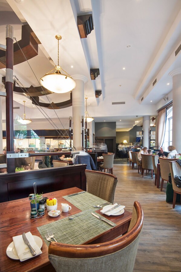 A spacious restaurant with high ceilings, modern lighting, and a central dining area with wooden tables and chairs. - The Commodore Hotel (Cape Town)
