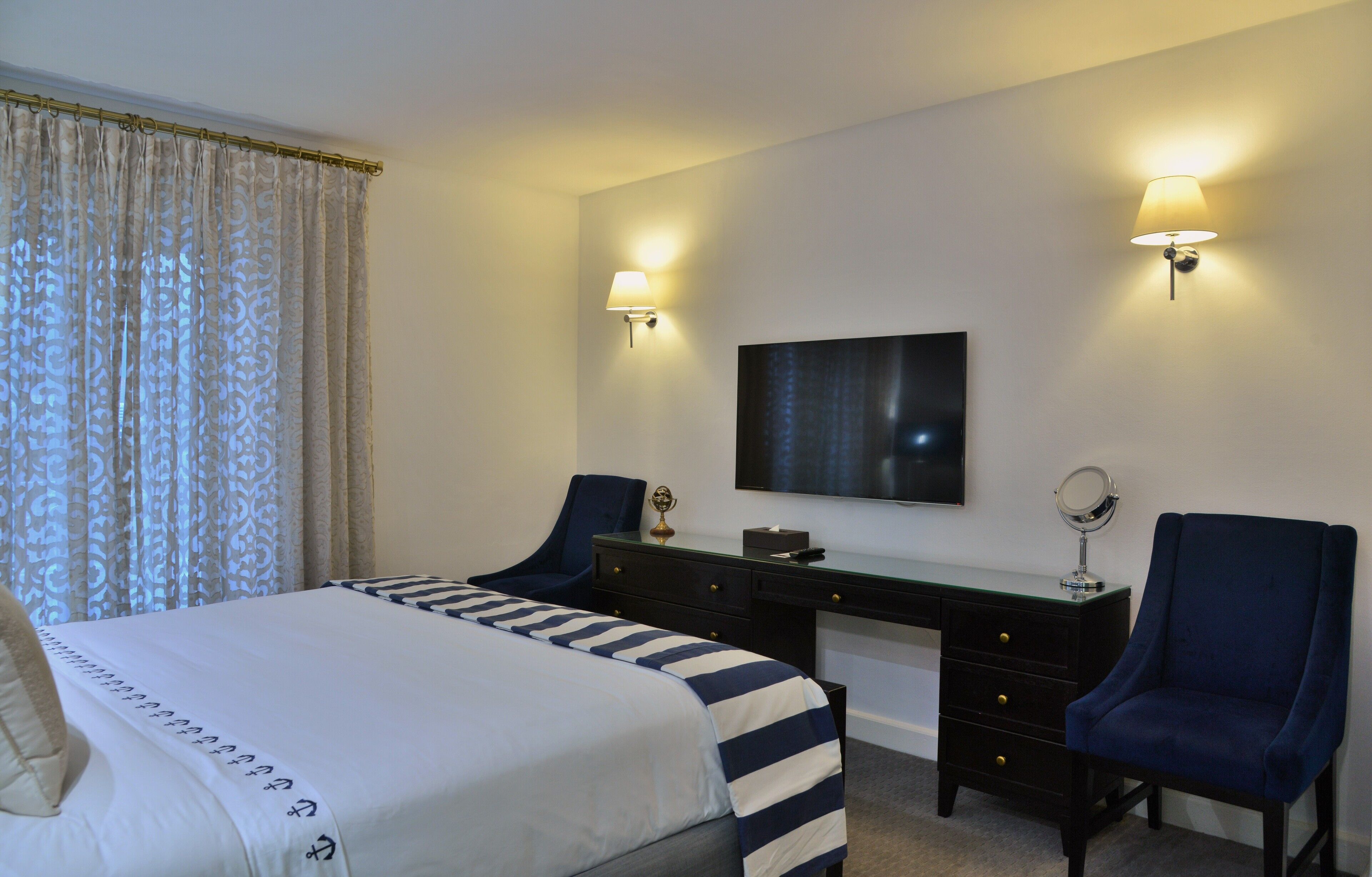 A hotel room with a bed, two chairs, a desk, a television, and two wall-mounted lamps.