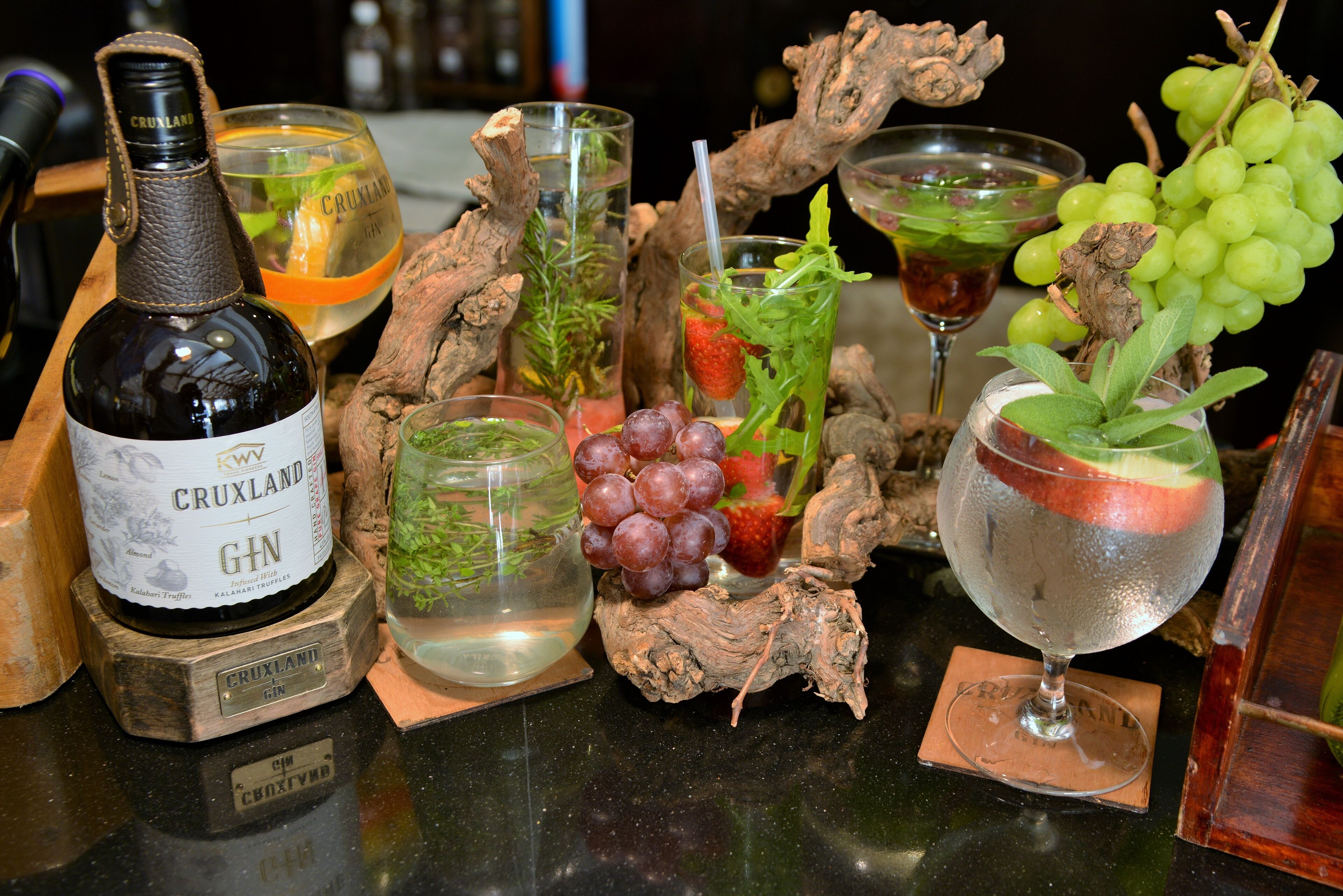 Eine Flasche Cruxland Gin, ein Glas mit Erdbeere und ein Glas mit grünen Trauben auf einer Bar.