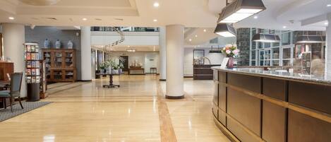 A spacious lobby with a polished wooden floor, a central staircase, and a reception desk with a vase of flowers.
