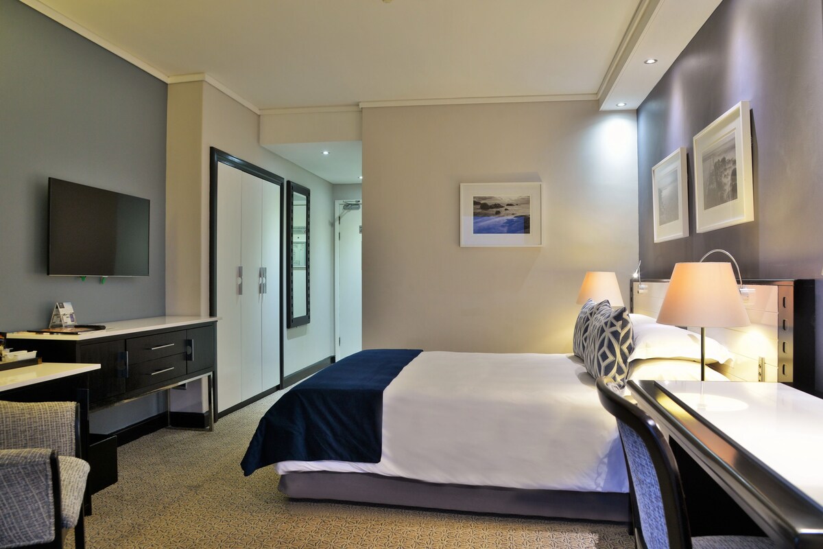 Habitación de hotel con cama, mesita de noche, lámpara, escritorio, silla y televisor en la pared.