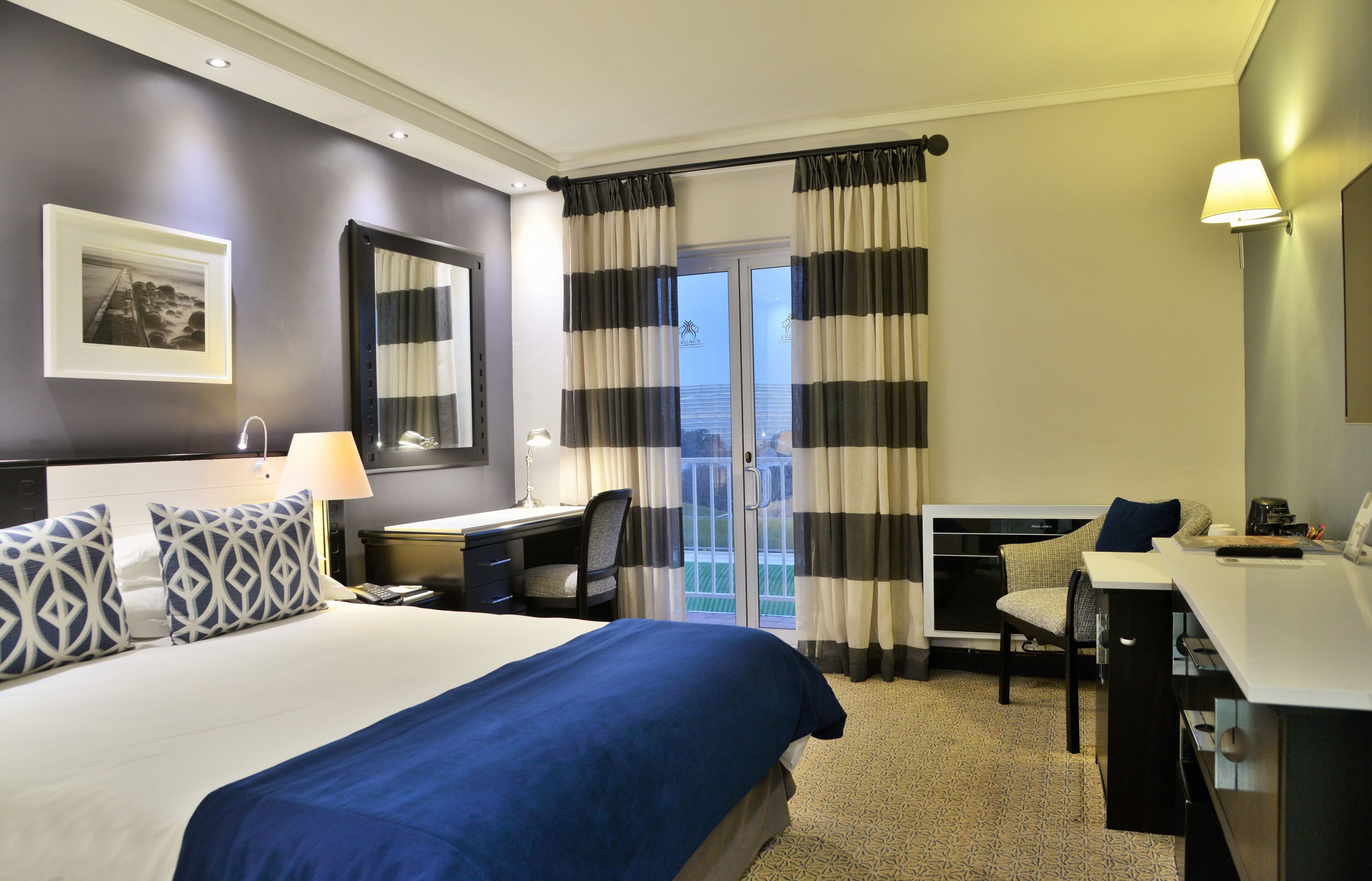 A hotel room with a large bed, a desk, a chair, a desk lamp, a telephone, and a window with curtains.