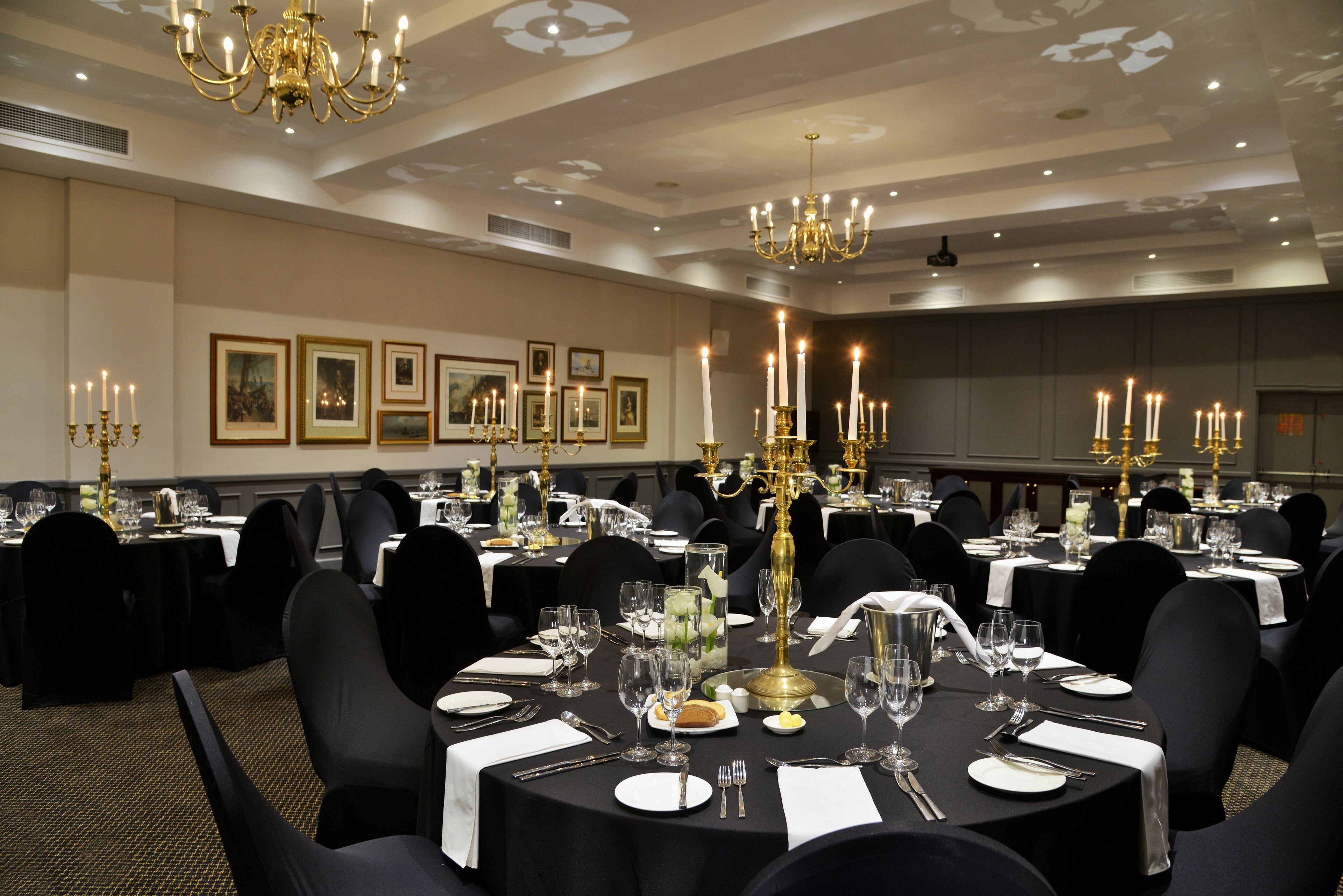 A banquet hall with round tables, black chairs, and chandeliers.