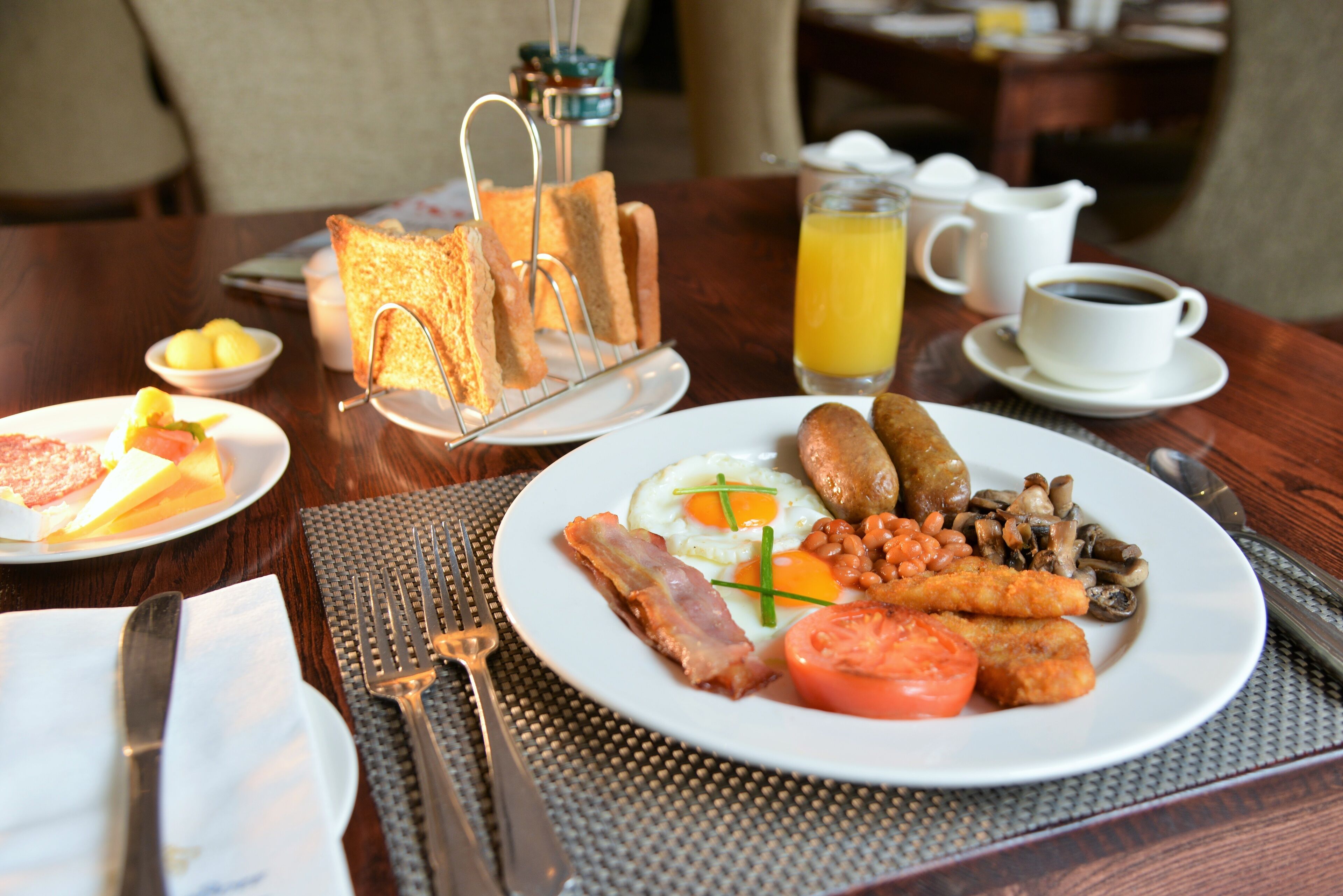 A breakfast setting with toast, eggs, sausages, beans, mushrooms, and tomatoes on a plate, accompanied by a glass of orange juice and a cup of coffee.