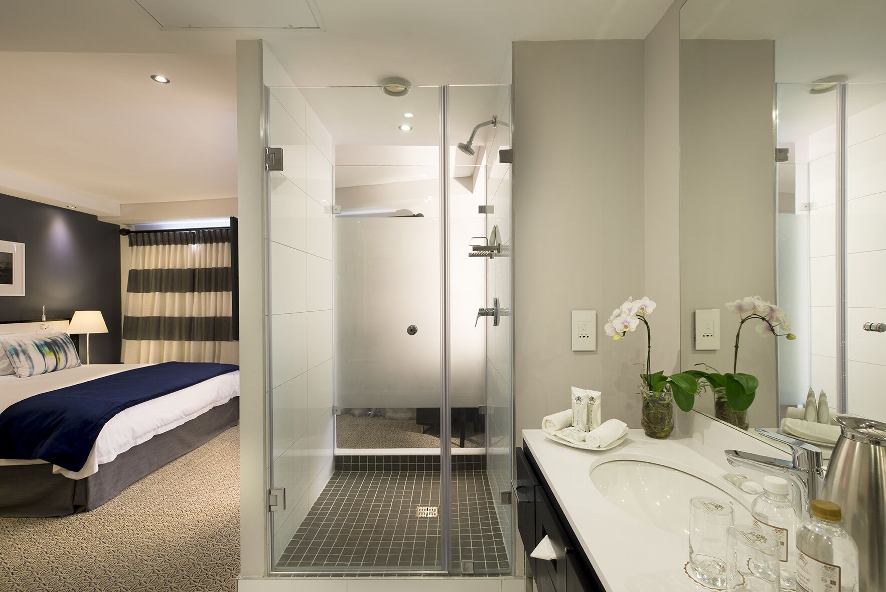A modern hotel room with a glass-enclosed shower, a large bed with a dark headboard, a white bathroom with a large mirror, and a small potted orchid on the vanity.