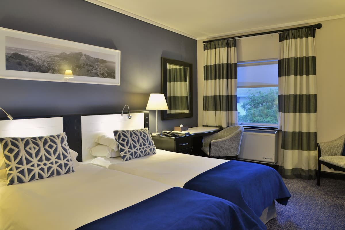 A hotel room with two beds, a desk, a chair, a lamp, a mirror, and a window with curtains.