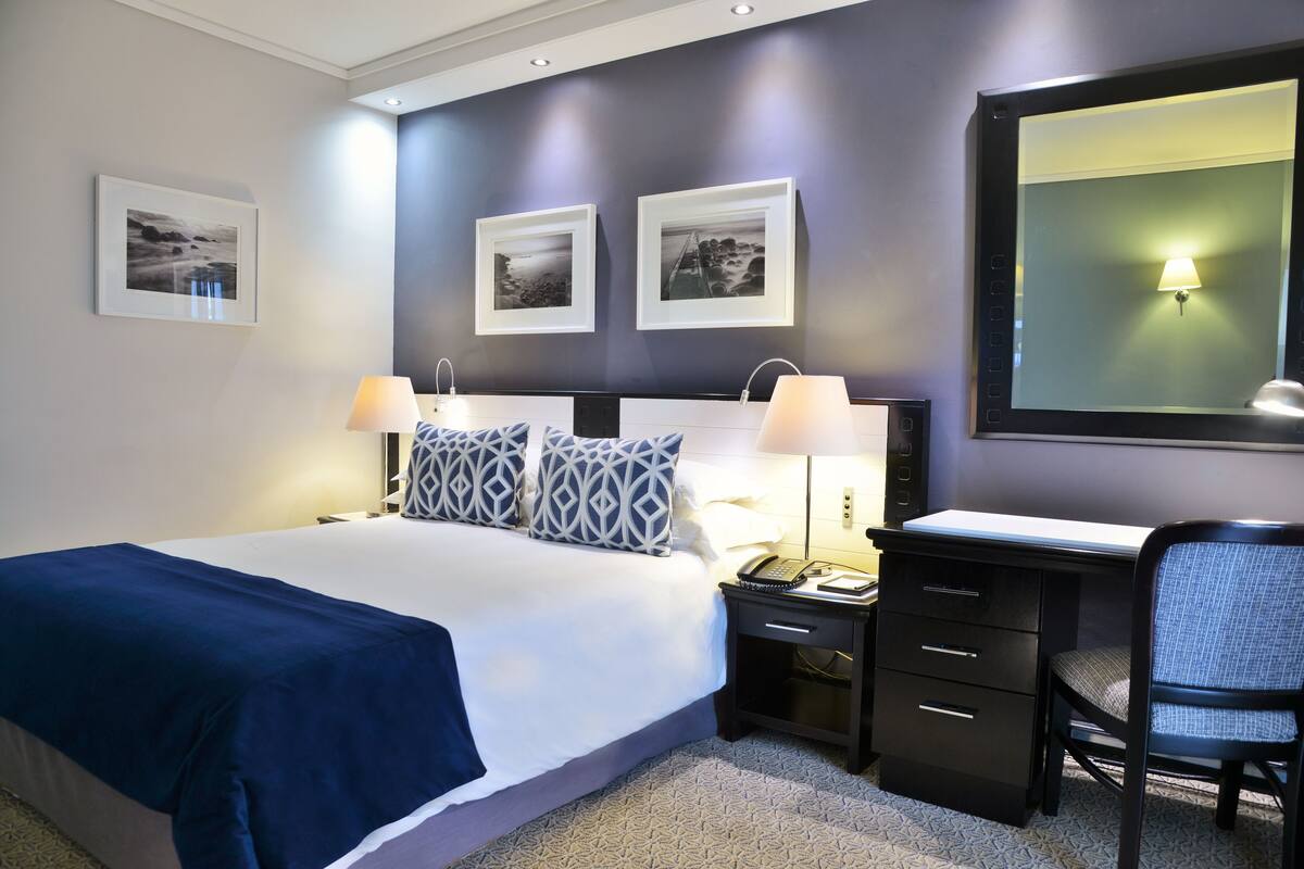 A hotel room with a bed, bedside tables, a desk, and a chair. There are framed pictures on the wall and a lamp on the desk.