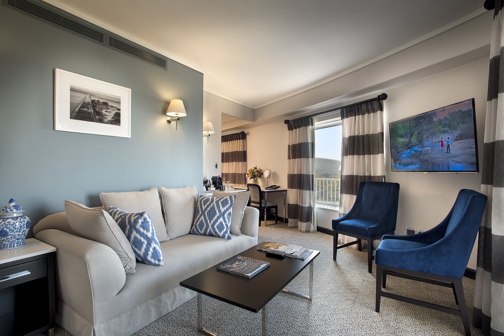 A modern hotel room with a sofa, armchairs, a coffee table, a TV, and a balcony with curtains.