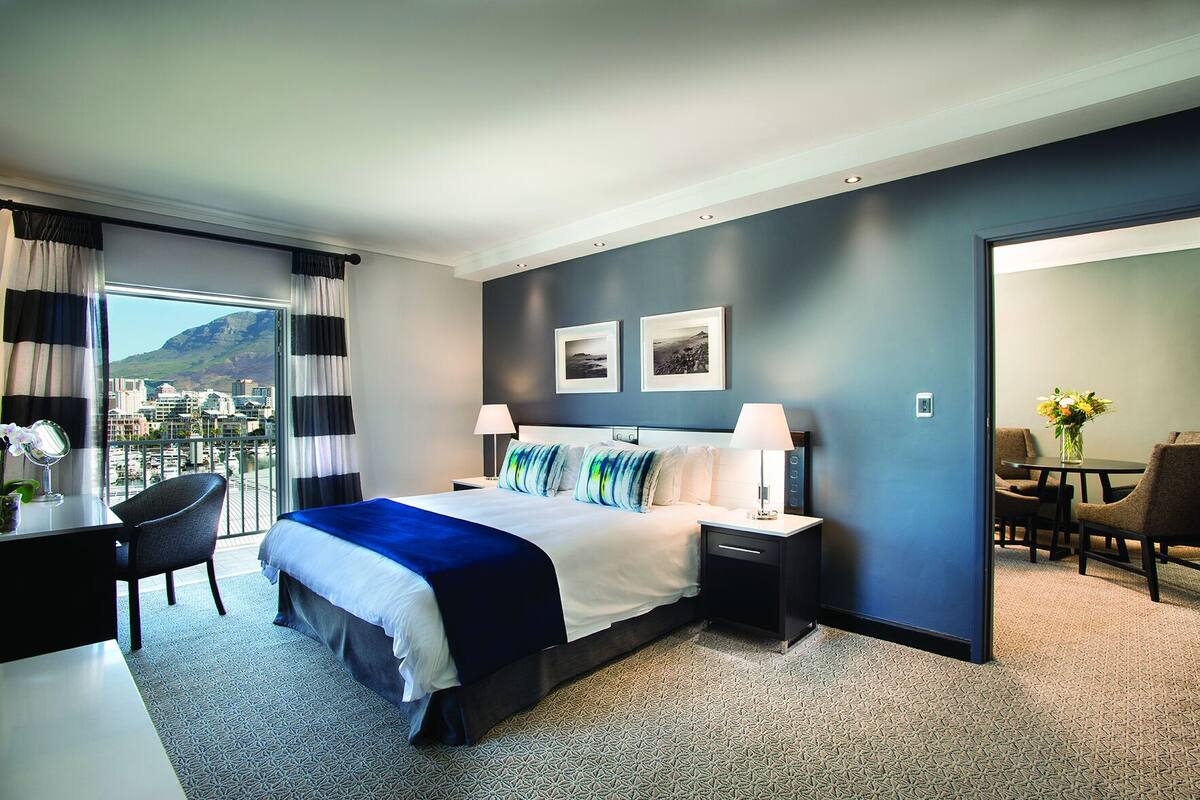 A hotel room with a large bed, a desk with a chair, a small table with chairs, and a view of a cityscape through a window.