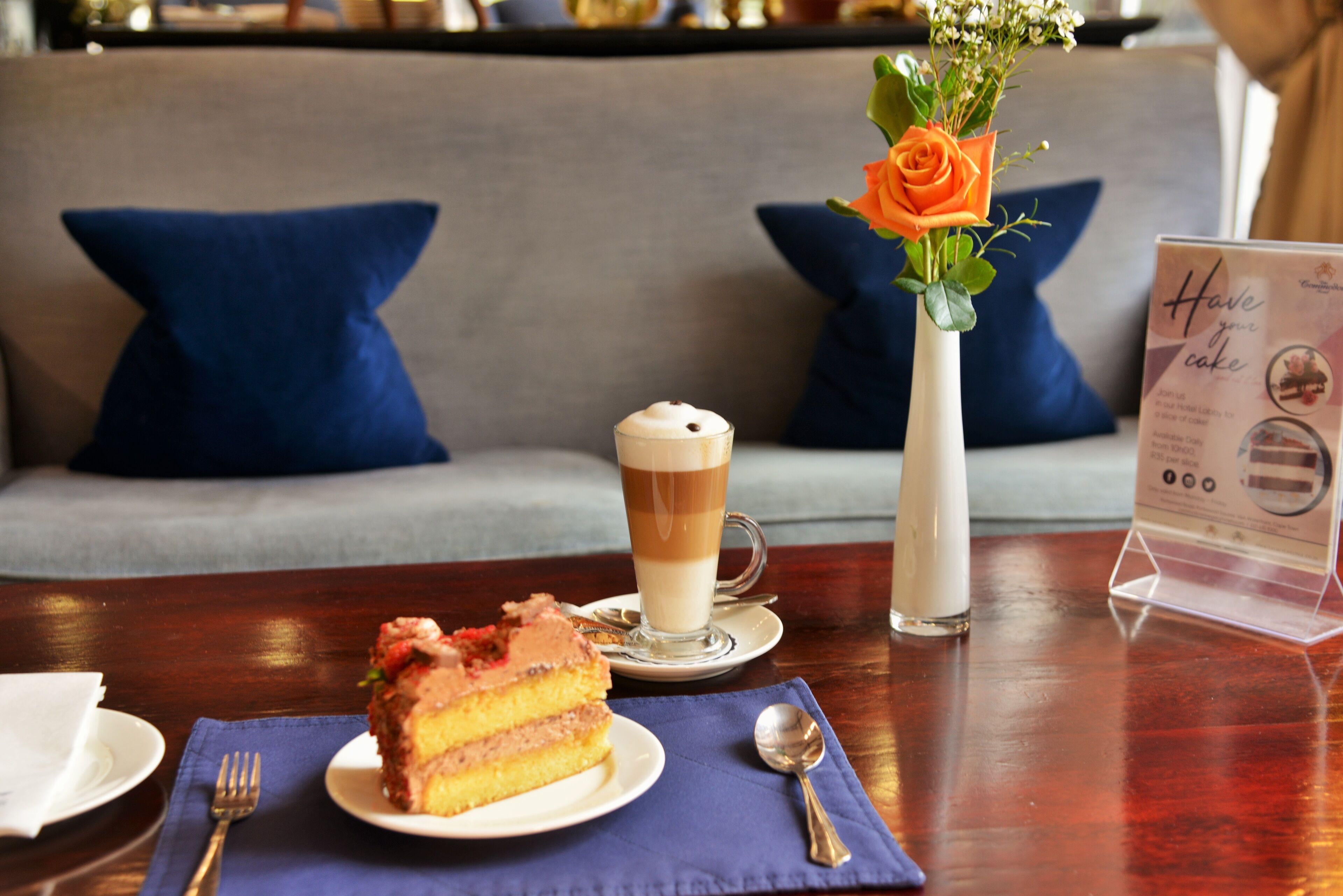A dining table with a slice of cake, a glass of coffee, and a vase with a rose.