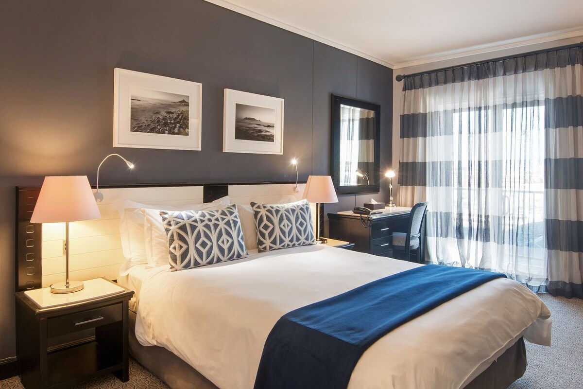 Habitación de hotel con una cama grande, dos mesitas de noche, un escritorio y una silla. Hay dos cuadros en la pared, una ventana con cortinas y una alfombra estampada.