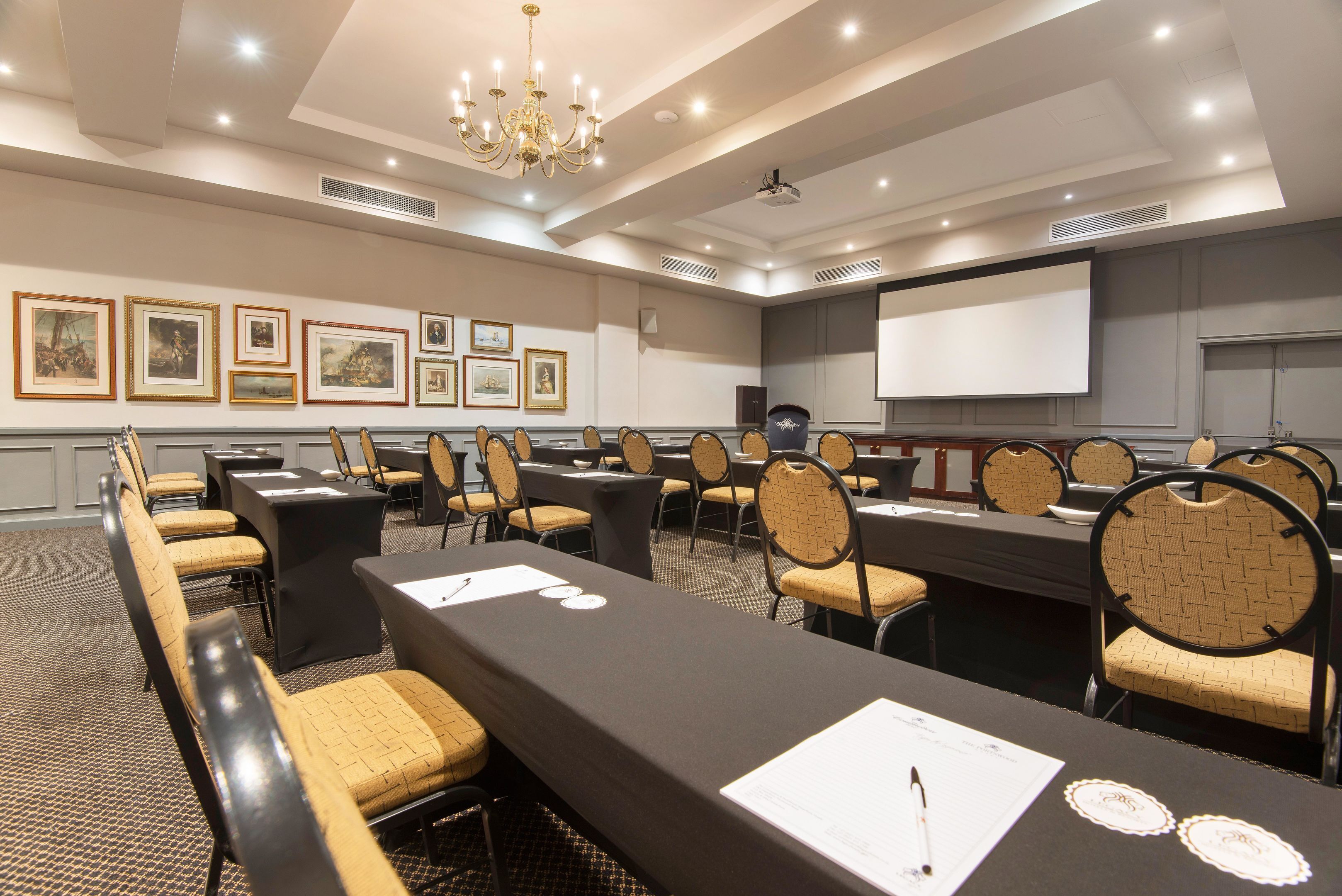A conference room with long tables, chairs, a projector screen, and framed artwork on the walls.