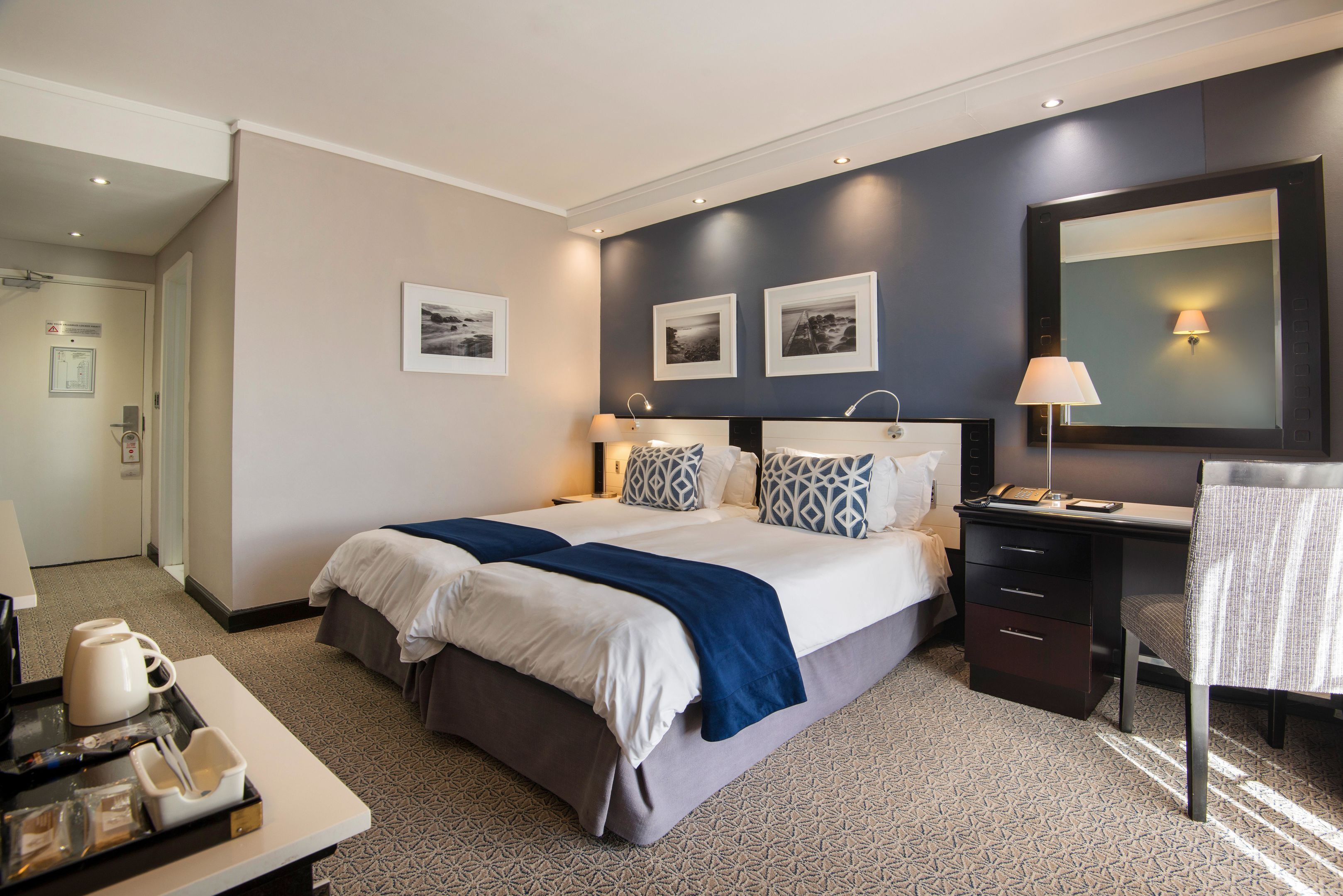 Une chambre d’hôtel avec un grand lit, des tables de chevet, un bureau et une chaise. Les murs sont ornés de tableaux encadrés, il y a une lampe et un plateau avec une cafetière et des tasses.