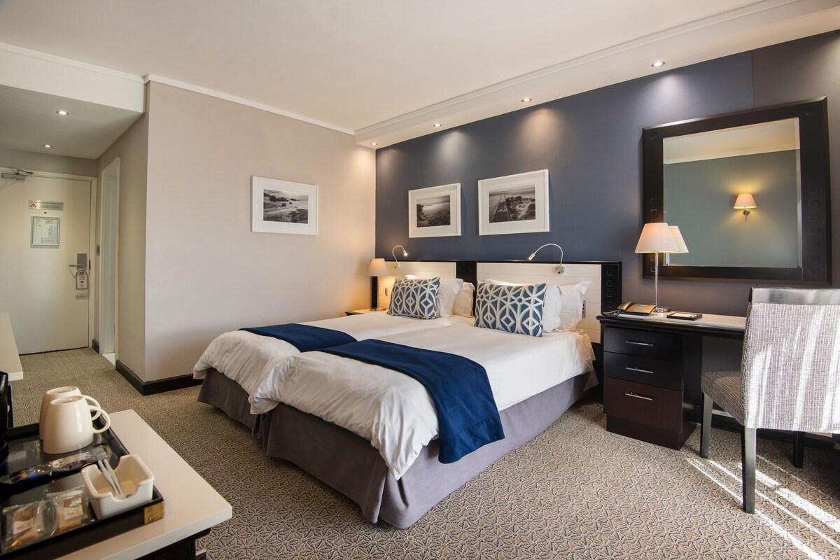 Habitación de hotel con una cama grande, mesitas de noche, un escritorio y una silla. En la pared hay cuadros enmarcados, una lámpara y una bandeja con una cafetera y tazas.