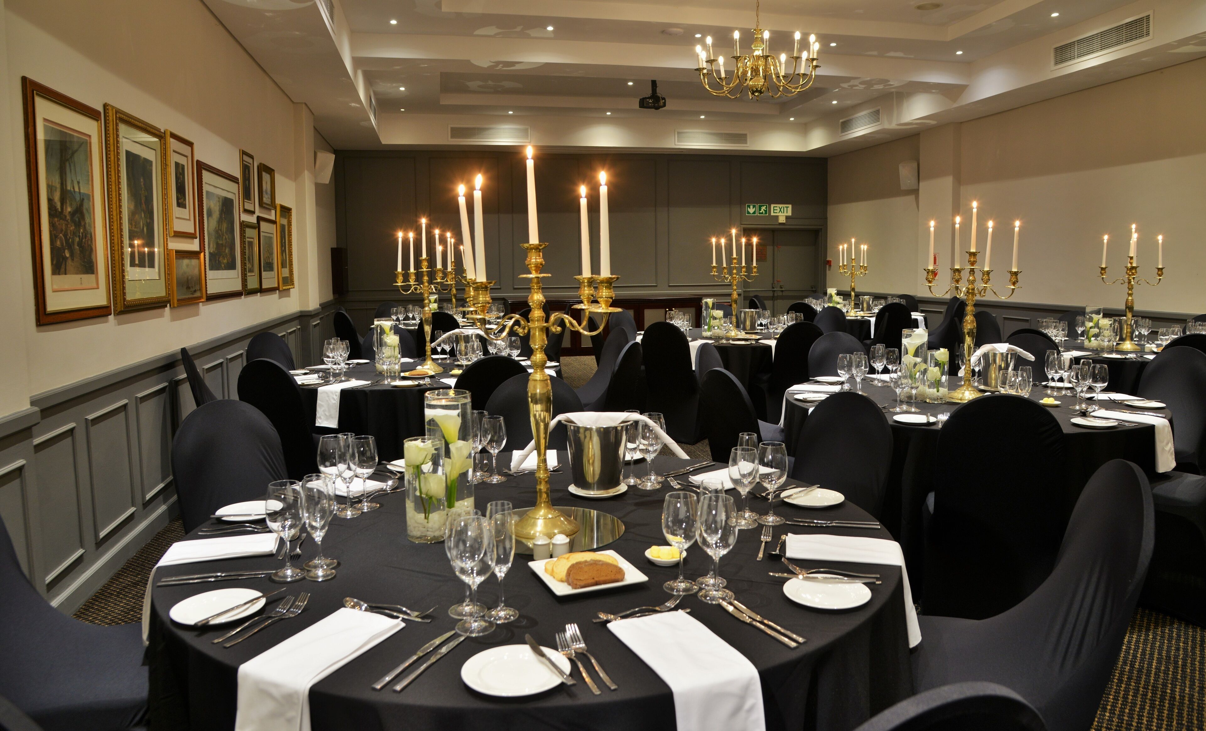 Een elegante eetzaal met ronde tafels gedekt voor een maaltijd, met gouden kandelaars, witte tafelkleden en zwarte stoelen.