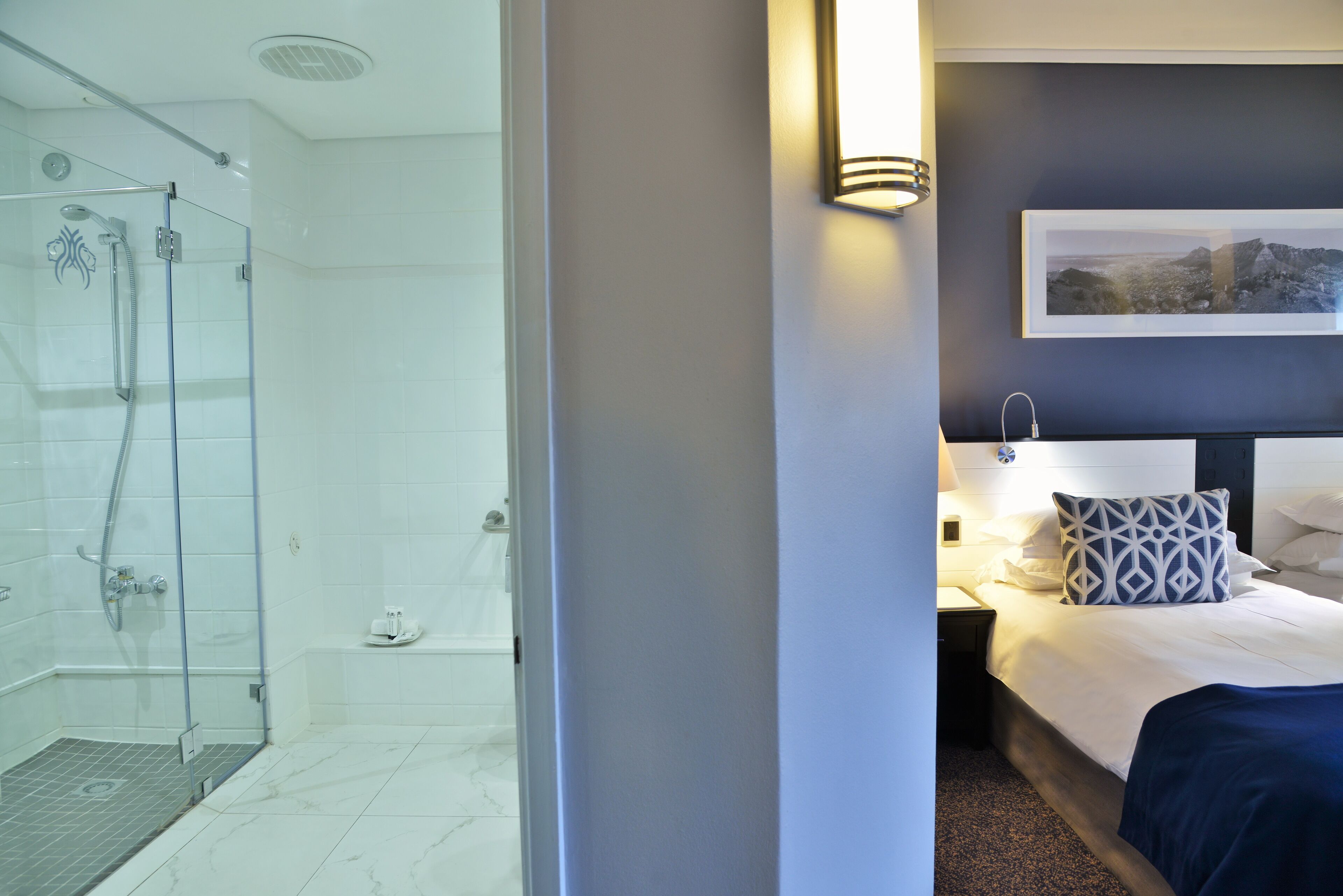 Ett hotellrum med en dusch som är omgiven av glasväggar, en säng med vita sängkläder och en geometrisk formad kudde samt en väggmonterad lampa.